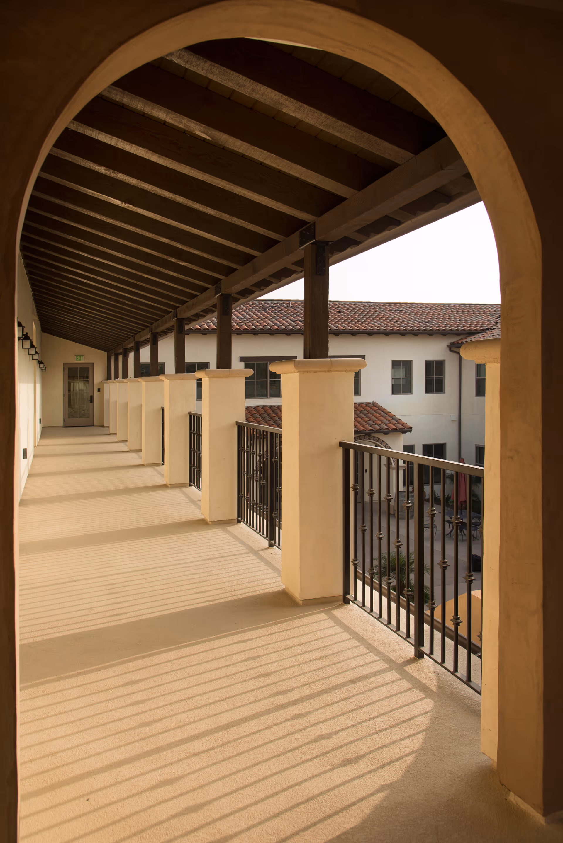 A covered outdoor corridor with a wooden beam ceiling and beige columns supporting a black metal railing, overlooking a courtyard with a building featuring white walls and red tile roofing.