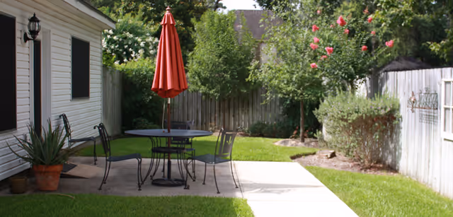 Outdoor patio with round table, chairs, red umbrella, and green lawn beside a white building.
