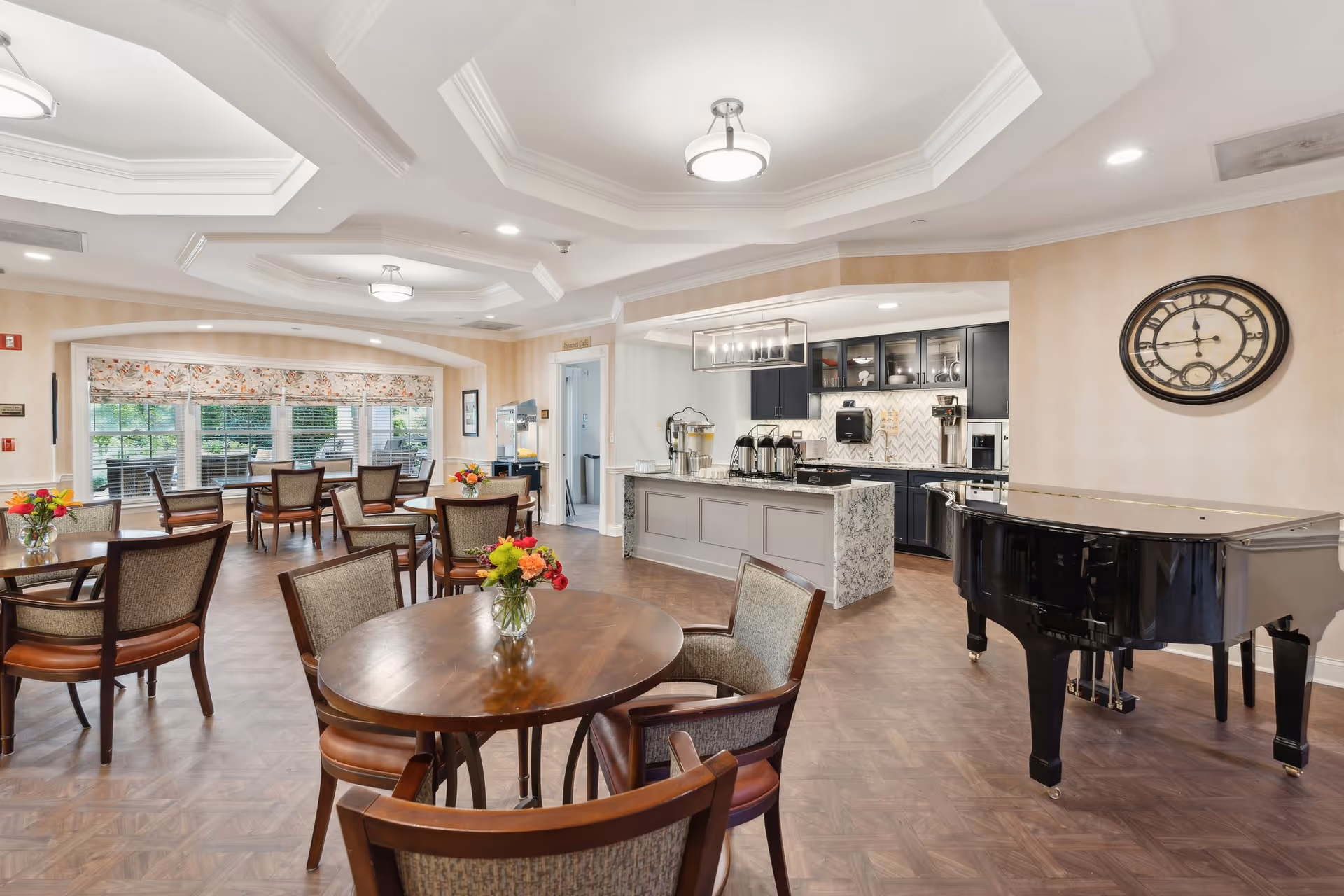 A spacious dining area in a senior living facility featuring multiple round wooden tables with chairs, each table adorned with a small vase of colorful flowers. In the background, there is a kitchen area with a large island, coffee dispensers, and cabinetry. A black grand piano is positioned to the right, and a large decorative wall clock hangs above it. The room has large windows with floral-patterned valances, recessed lighting, and a coffered ceiling.