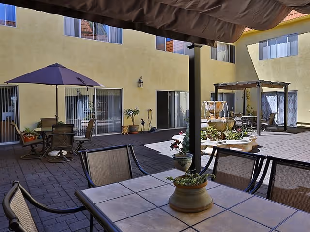Outdoor courtyard area at Villa Redondo with a tiled table and chairs in the foreground, potted plants on the table, a large umbrella shading another table with chairs, a multi-tiered water fountain, and yellow building walls with windows and sliding glass doors surrounding the courtyard.