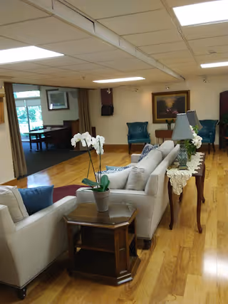 A spacious living room area with wooden flooring, featuring a beige sofa and armchair with blue cushions, a wooden side table with a potted white orchid, and a long wooden console table with a lace runner and two lamps. In the background, there are two teal armchairs, a framed painting on the wall, and a dining area visible through an open doorway.