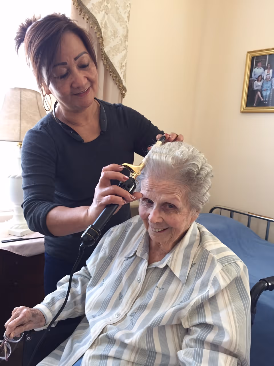 A woman is curling the hair of an elderly woman who is sitting in a wheelchair and smiling. They are in a cozy room with a bed, a lamp, and a framed family photo on the wall.
