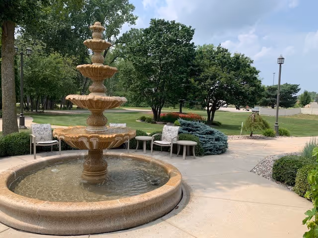 A multi-tiered stone fountain in a landscaped outdoor courtyard with chairs, trees, and walking paths.