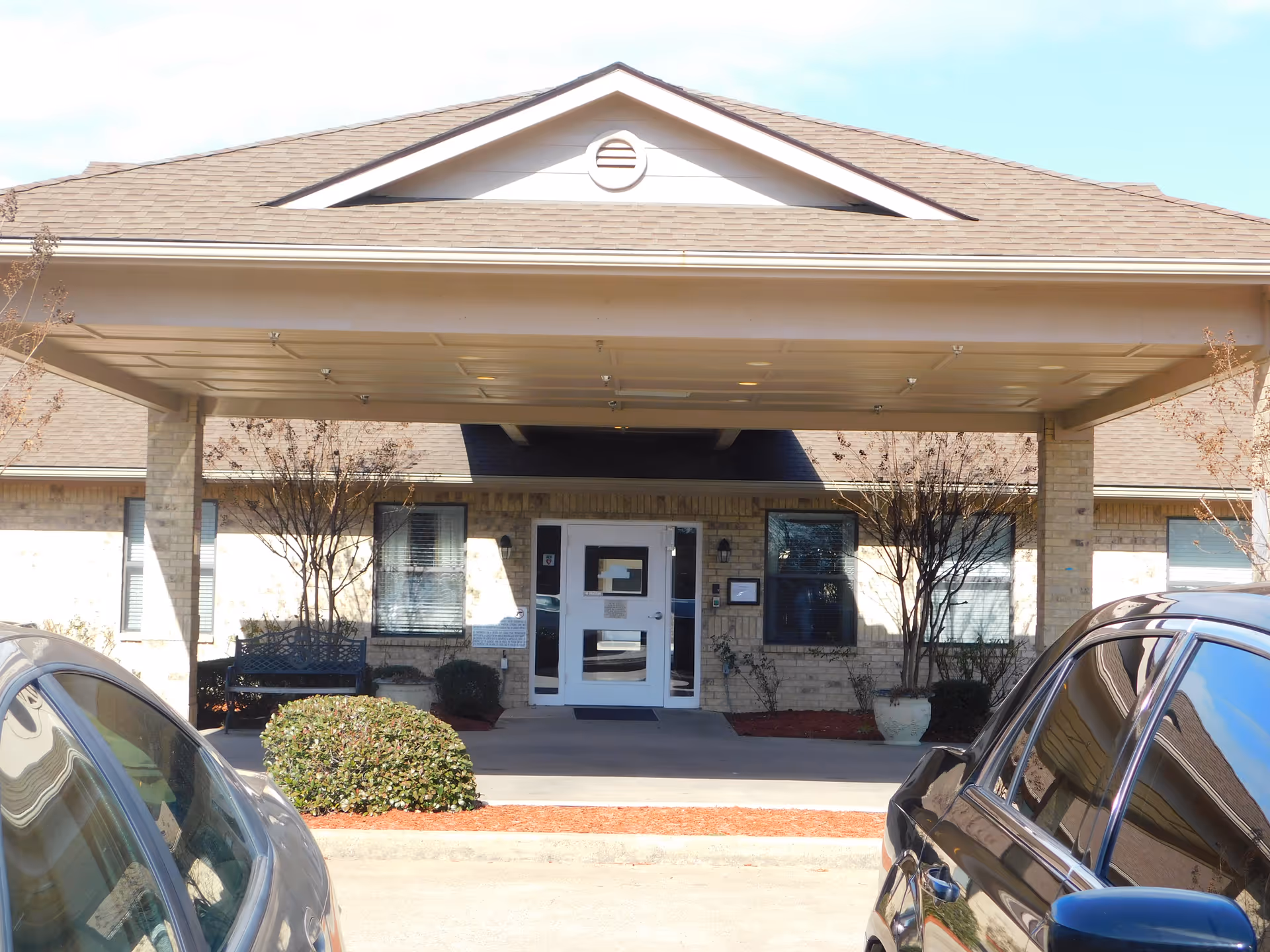 Front entrance of Fairfield Nursing and Rehabilitation Center showing a covered drop-off area with two parked cars in front, beige brick exterior walls, windows with blinds, and some landscaping including bushes and small trees.