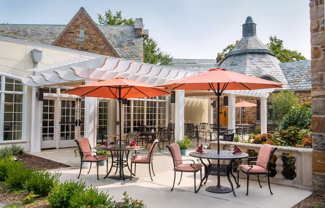 Outdoor patio area at Winchester Gardens featuring several round tables with pink umbrellas and cushioned chairs. The patio is adjacent to a stone building with large windows and a white pergola. There are plants and greenery surrounding the patio.