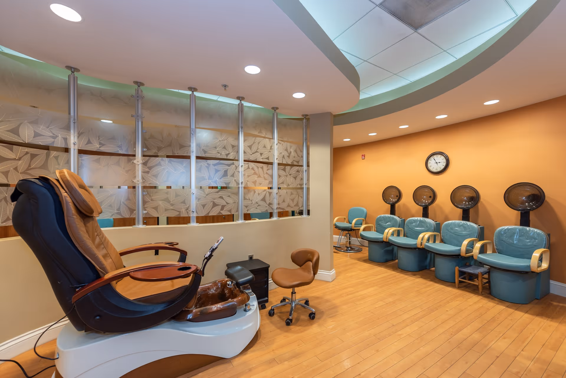 Interior view of a salon area with a pedicure chair and foot bath in the foreground, and a row of teal salon chairs with hair dryers against an orange wall in the background. The floor is wooden, and there is a frosted glass partition with a leaf pattern separating the space.