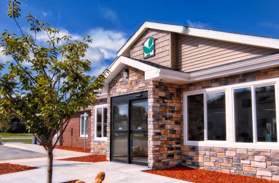 Front entrance of a single-story assisted living building with a stone facade, glass double doors, large windows, and a small tree in the foreground.