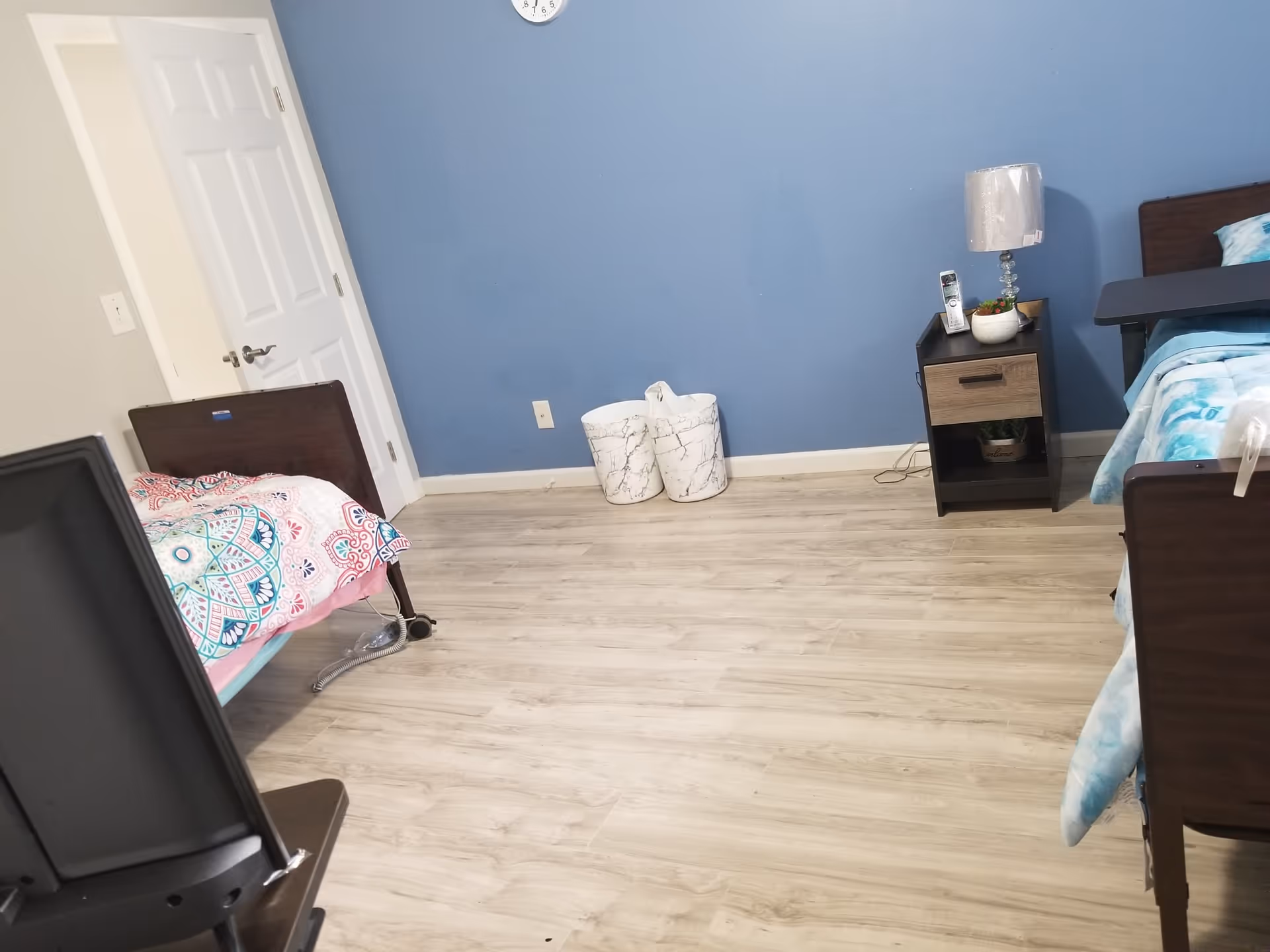 A room with two single beds on either side, each with colorful bedding. There is a small nightstand between the beds with a lamp, a phone, and a small plant. The walls are painted blue and white, and there is a clock on the blue wall. The floor is light wood, and there are two white marble-patterned waste bins against the blue wall.