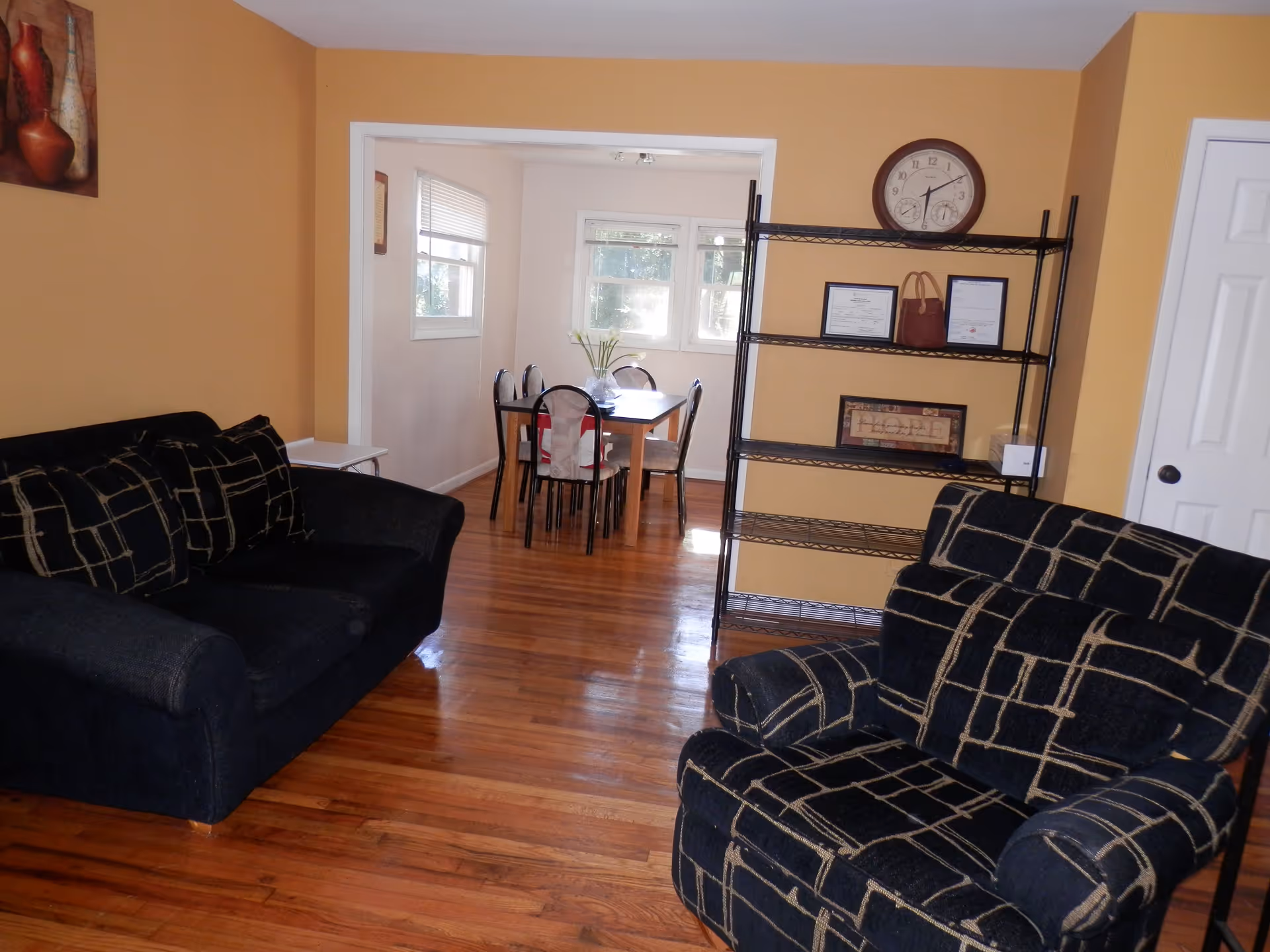 Interior view of a living room with two black patterned couches, a metal shelving unit with a clock and framed items, and a dining area with a table and chairs visible through an open doorway. The walls are painted a warm yellow and the floor is wooden.