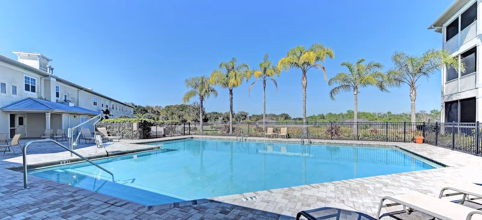 Outdoor swimming pool and deck with lounge chairs and palm trees beside a senior living building under a clear blue sky.
