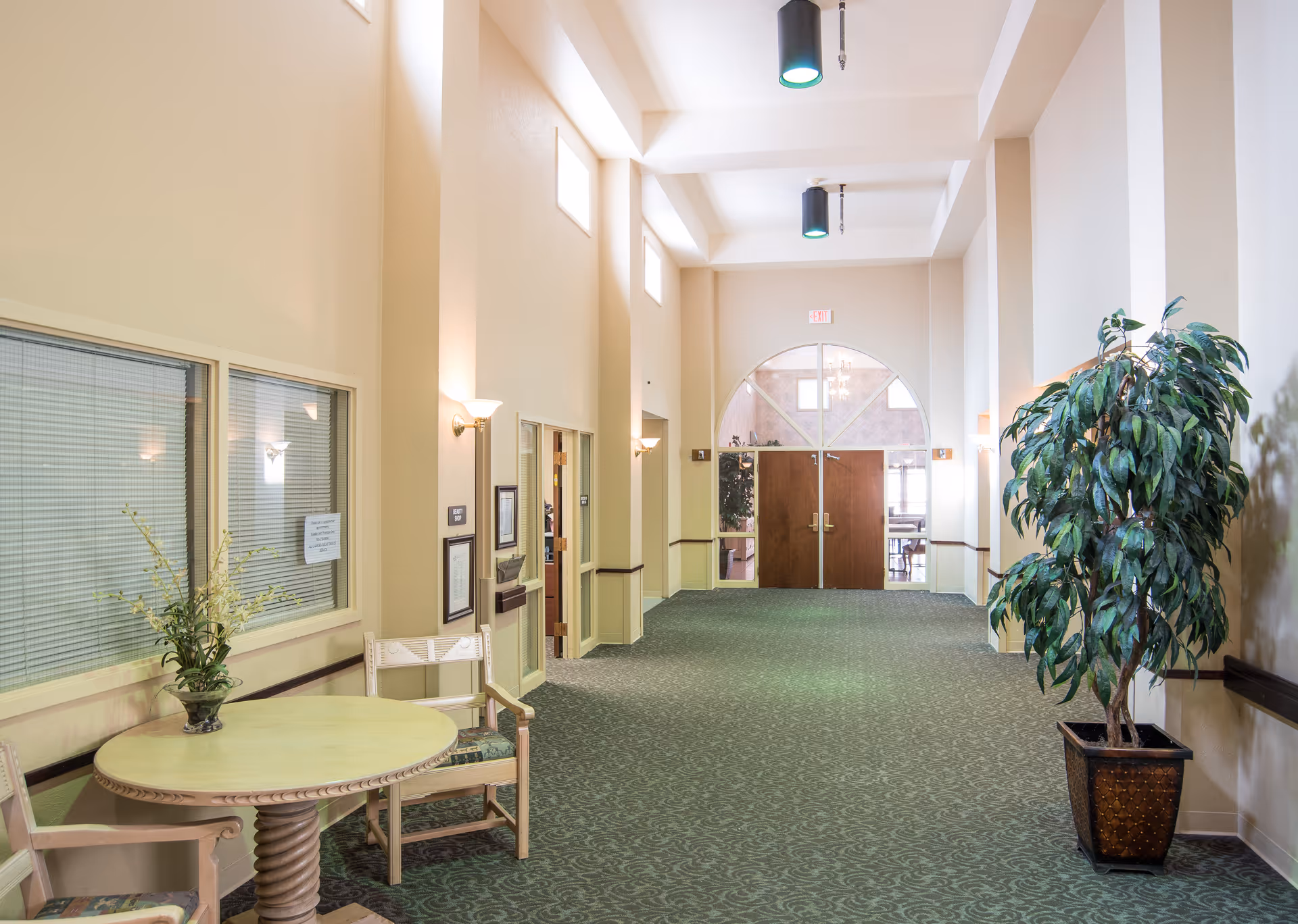 A spacious, well-lit hallway with high ceilings and green patterned carpet. There is a round table with two chairs and a vase with flowers on the left side, and a large potted plant on the right side. The hallway leads to double wooden doors with glass panels and an arched window above them. Wall sconces provide additional lighting along the walls.