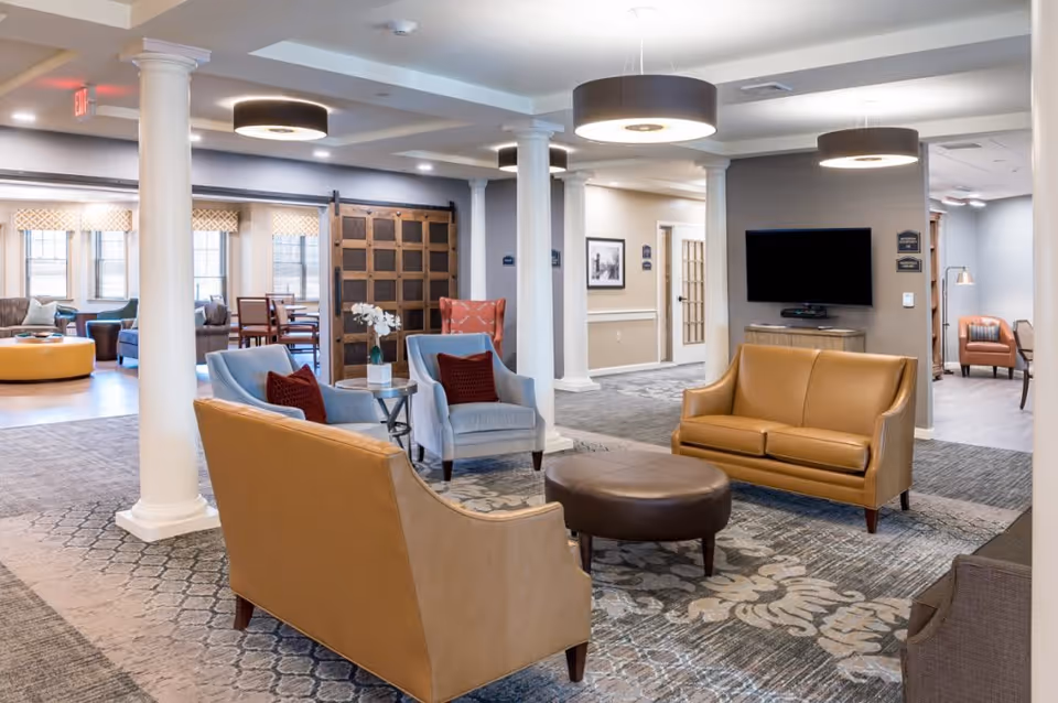 A spacious and well-lit living room area in Arbor Garden Place featuring comfortable seating including tan leather sofas, blue armchairs with red cushions, a round brown ottoman, and a patterned carpet. The room has white columns, modern ceiling lights, a wall-mounted flat-screen TV, and large windows letting in natural light. In the background, there is a dining area with tables and chairs.