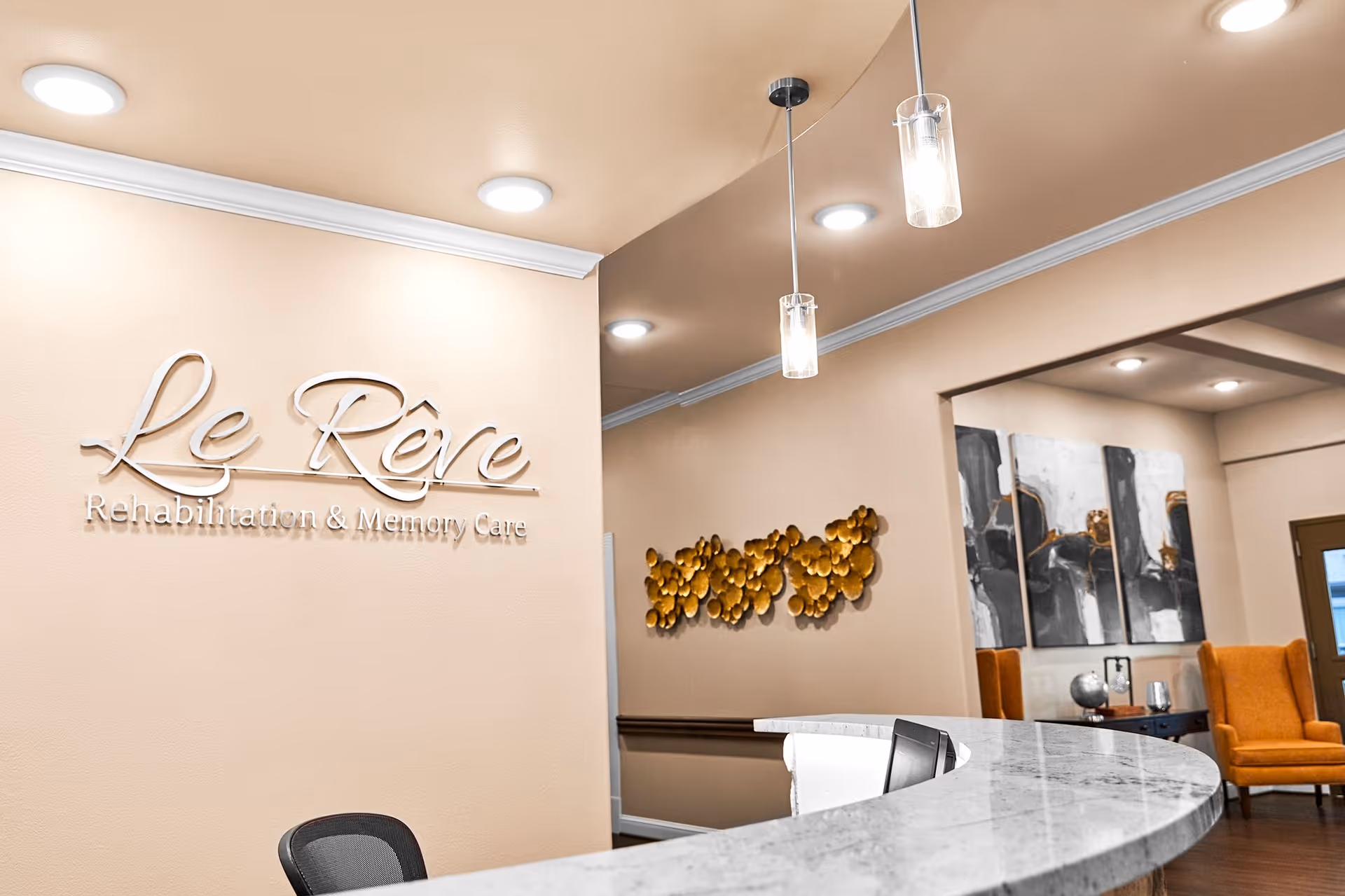 Reception area of Le Rêve Rehabilitation and Memory Care center featuring a curved marble countertop, a black office chair, beige walls with the facility's name in metallic letters, modern pendant lights, decorative wall art, and orange armchairs in the background.