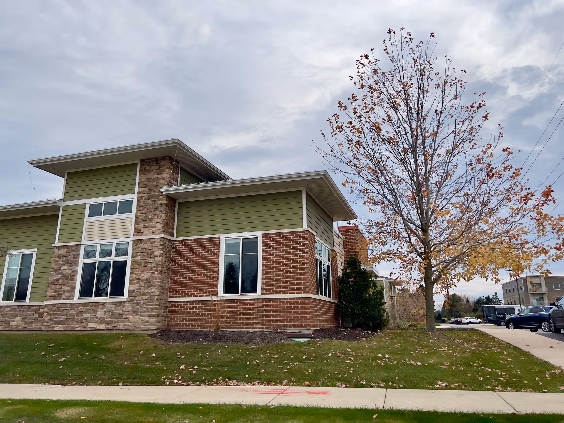 Exterior view of a modern building with a combination of stone, brick, and green siding. There are several windows and a tree with autumn leaves on the right side. A sidewalk and parked cars are visible in the background under a cloudy sky.