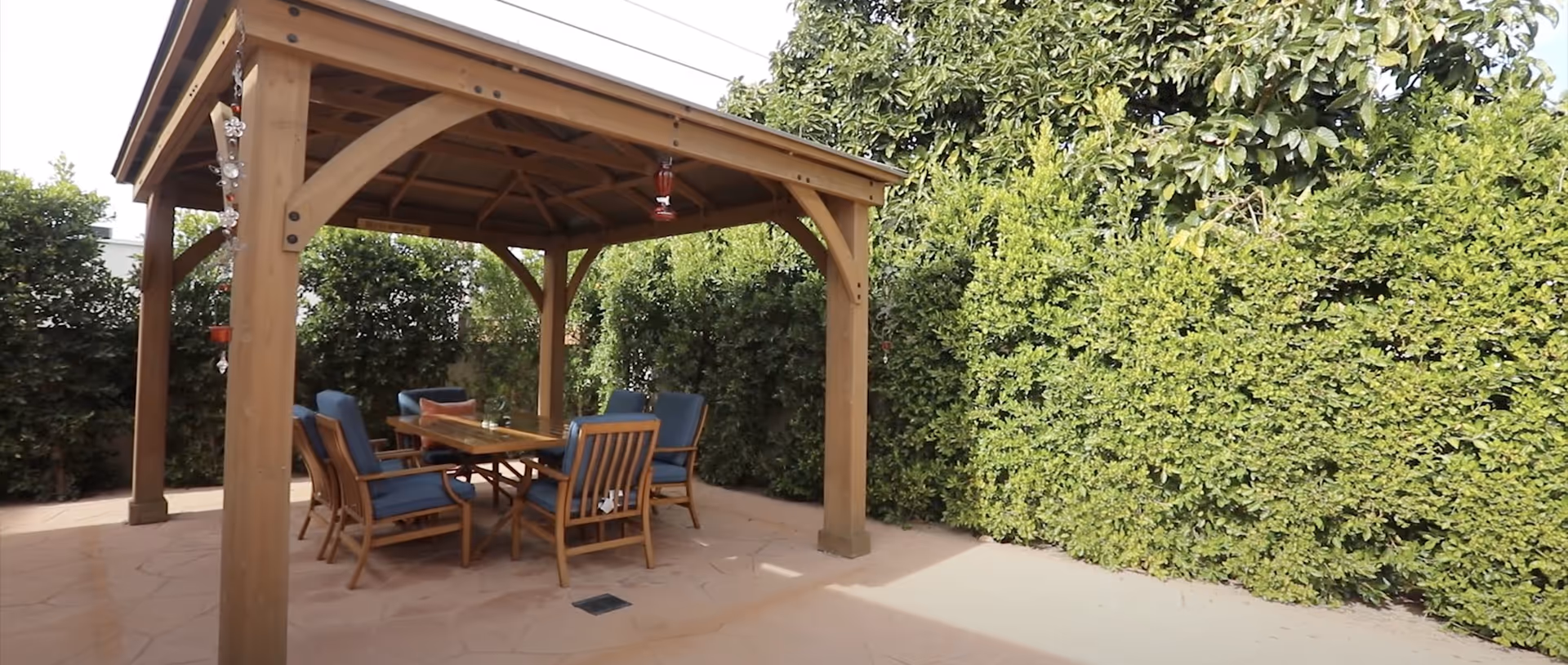 Outdoor wooden gazebo with a table and six cushioned chairs underneath, surrounded by tall green hedges and trees.
