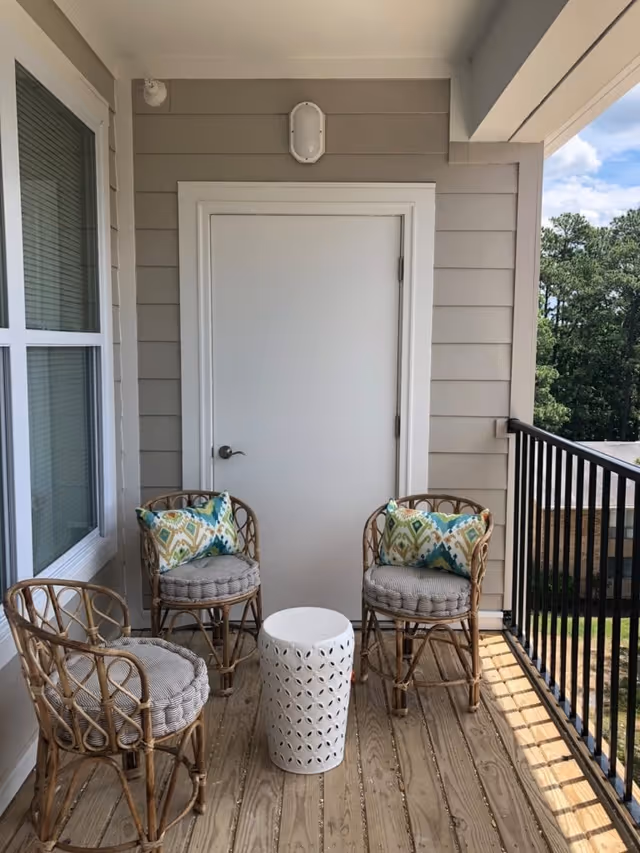 A small outdoor balcony area with three wicker chairs, two of which have patterned cushions, and a white decorative cylindrical side table. The balcony has wooden flooring, beige siding walls, a white door, a wall-mounted light fixture above the door, and a black metal railing. Trees and a building are visible in the background.