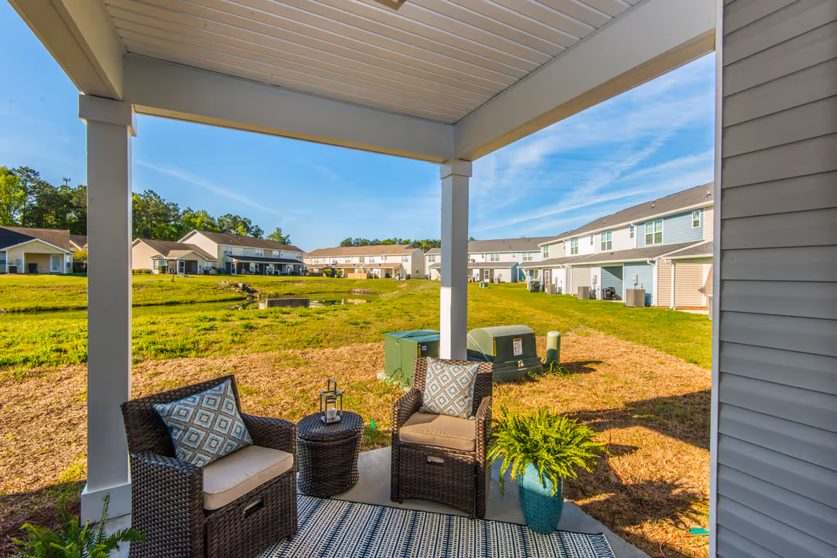 Covered back patio with wicker seating, small table and potted plant overlooking a grassy courtyard and rows of townhome-style housing.