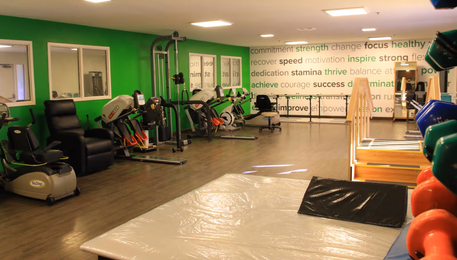 A rehabilitation or fitness room with exercise equipment including stationary bikes, a NuStep machine, resistance machines, a padded mat on the floor, and a wooden ramp with handrails. The walls are painted green and white, with motivational words like commitment, strength, focus, healthy, recover, speed, motivation, inspire, strong, dedication, stamina, thrive, balance, achieve, courage, success, determination, improve, and empower displayed on one wall.