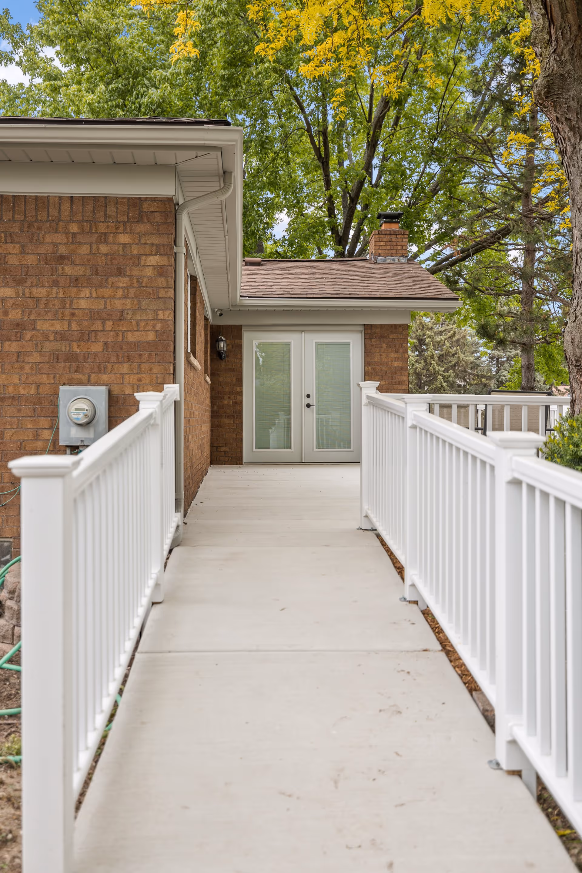 A concrete wheelchair-accessible ramp with white railings leading to a set of double glass doors on a brick building, surrounded by green trees.