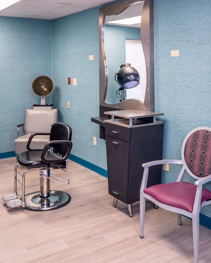Interior of a salon area in a nursing and rehabilitation center featuring a black salon chair with footrests, a gray chair with a hair dryer hood, a black cabinet with drawers and a large mirror above it, and a purple cushioned chair with a patterned backrest. The walls are teal with a textured finish and the floor is light wood.