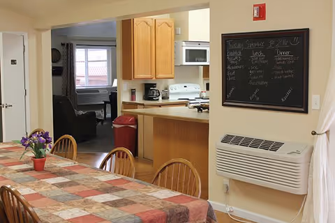 Interior view of a senior living facility dining area with a long table covered by a patchwork tablecloth and wooden chairs around it. A small potted plant with purple flowers is on the table. In the background, there is a kitchen area with wooden cabinets, a white stove, microwave, and a coffee maker. A chalkboard menu is mounted on the wall above a white heating/cooling unit. A living room with a window and armchair is visible through an open doorway.