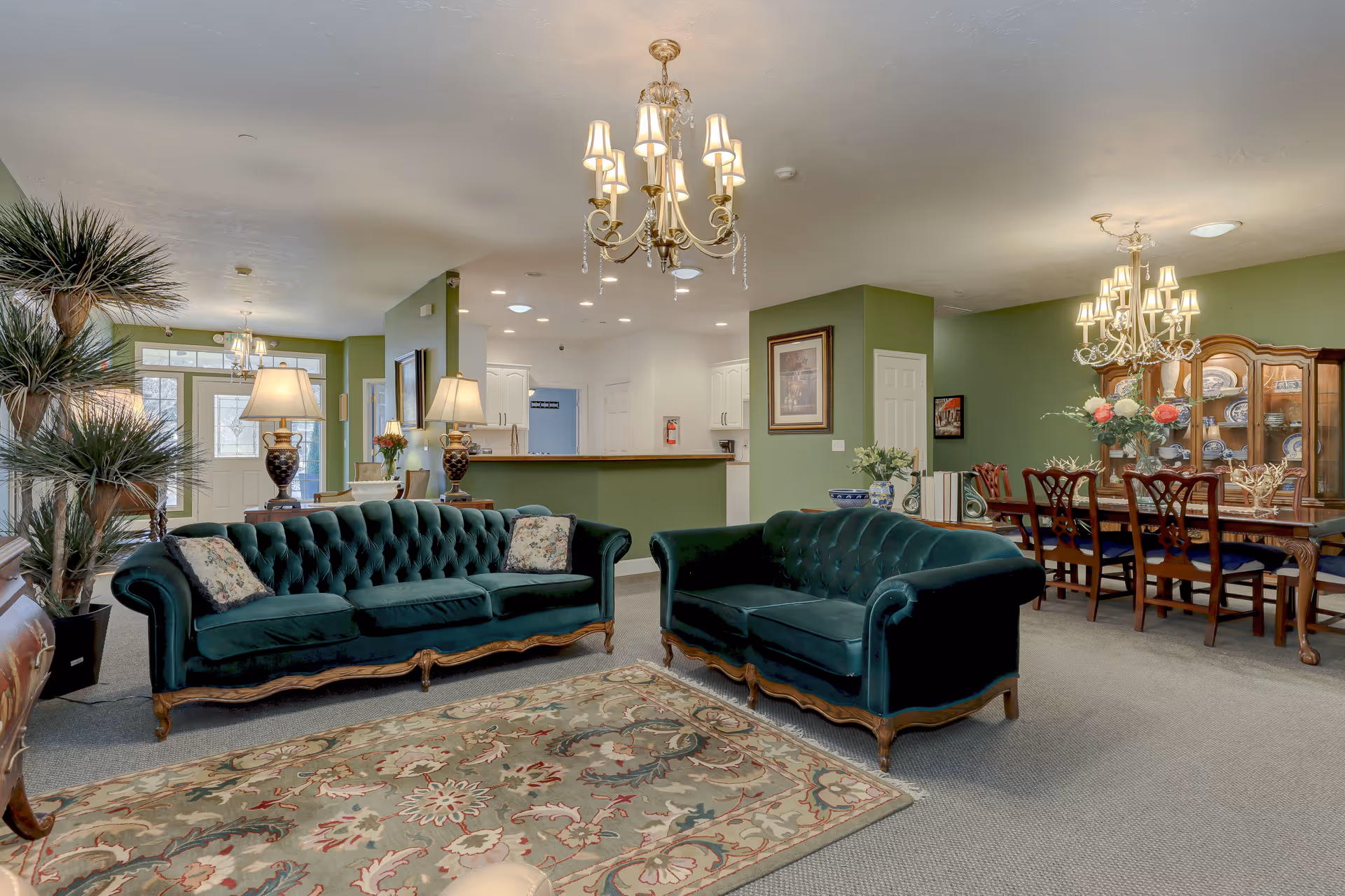 Spacious assisted-living common room with matching green velvet sofas, chandeliers, a dining table, and a patterned rug.