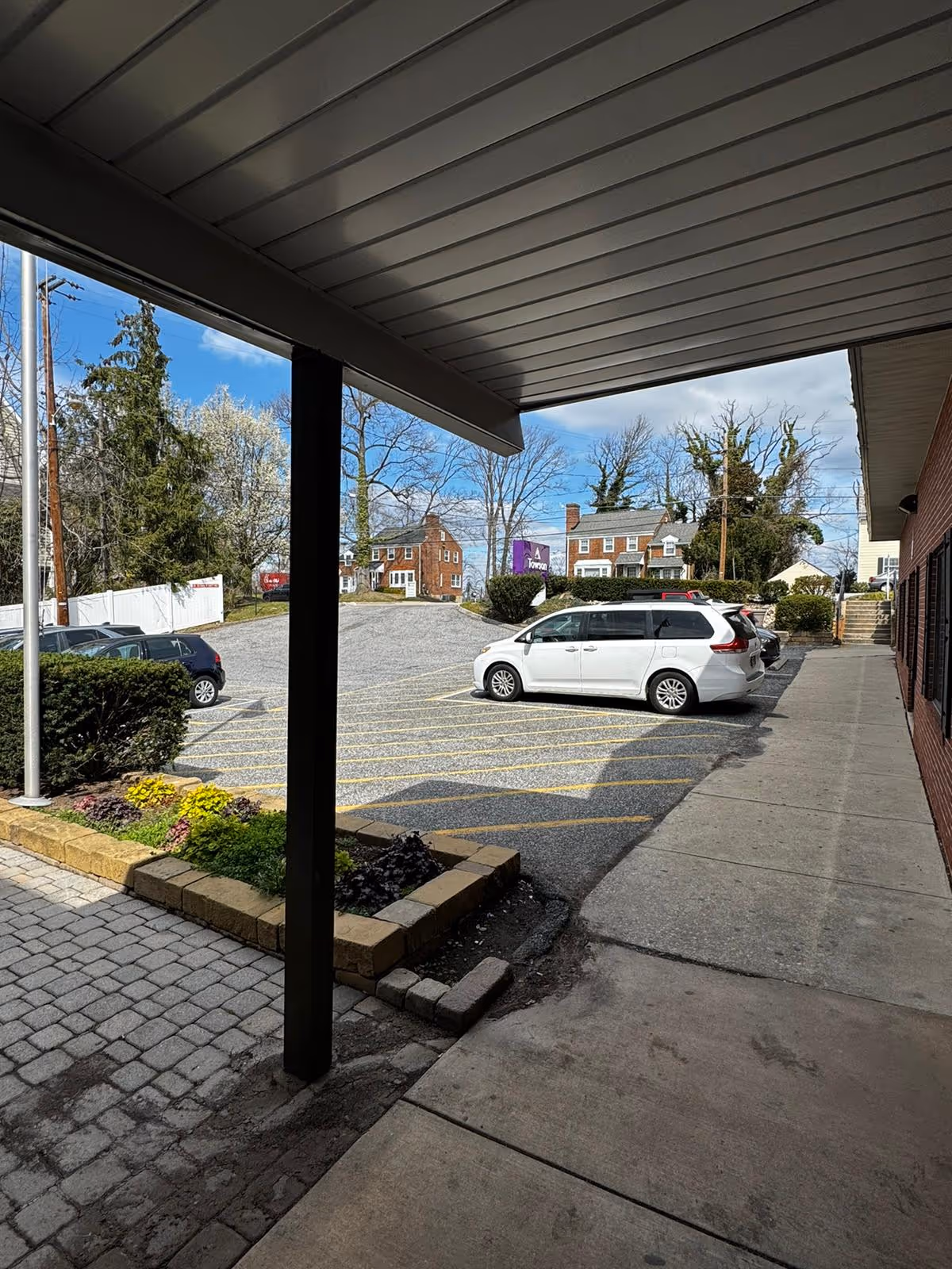 View from under a covered walkway looking out onto a parking lot with several parked cars, a small landscaped area with flowers and bushes, and residential houses in the background under a partly cloudy sky.