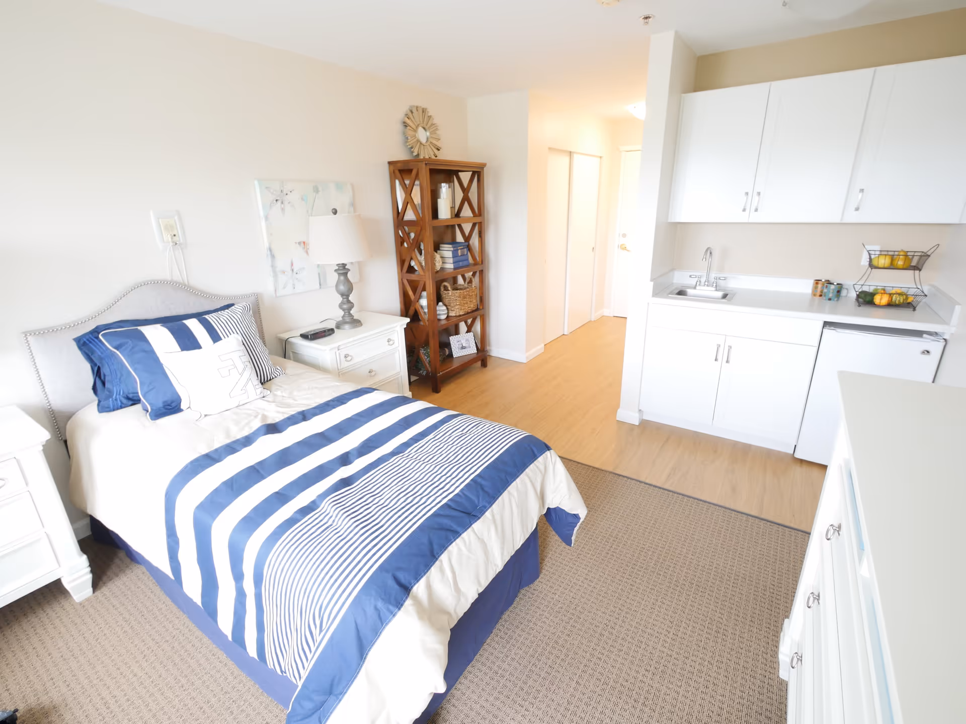 A bright and clean bedroom with a single bed dressed in blue and white striped bedding. Next to the bed is a white nightstand with a lamp and an alarm clock. There is a wooden shelving unit with decorative items and books. The room also features a small kitchenette with white cabinets, a sink, a mini fridge, and a two-tier fruit basket. The floor is a combination of carpet and wood, and the walls are painted white.