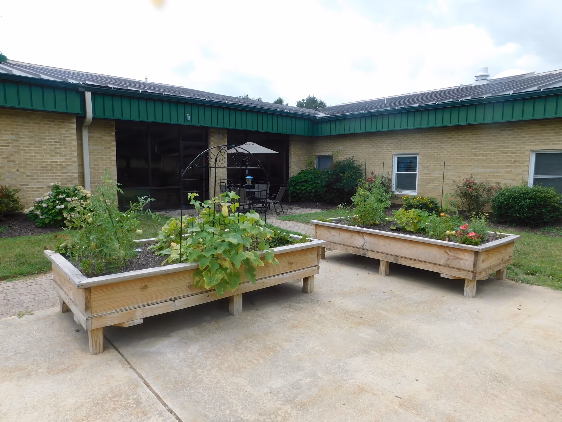 Raised wooden garden beds with plants on a concrete patio in front of a single-story brick senior living building with green trim and a patio table under an umbrella.