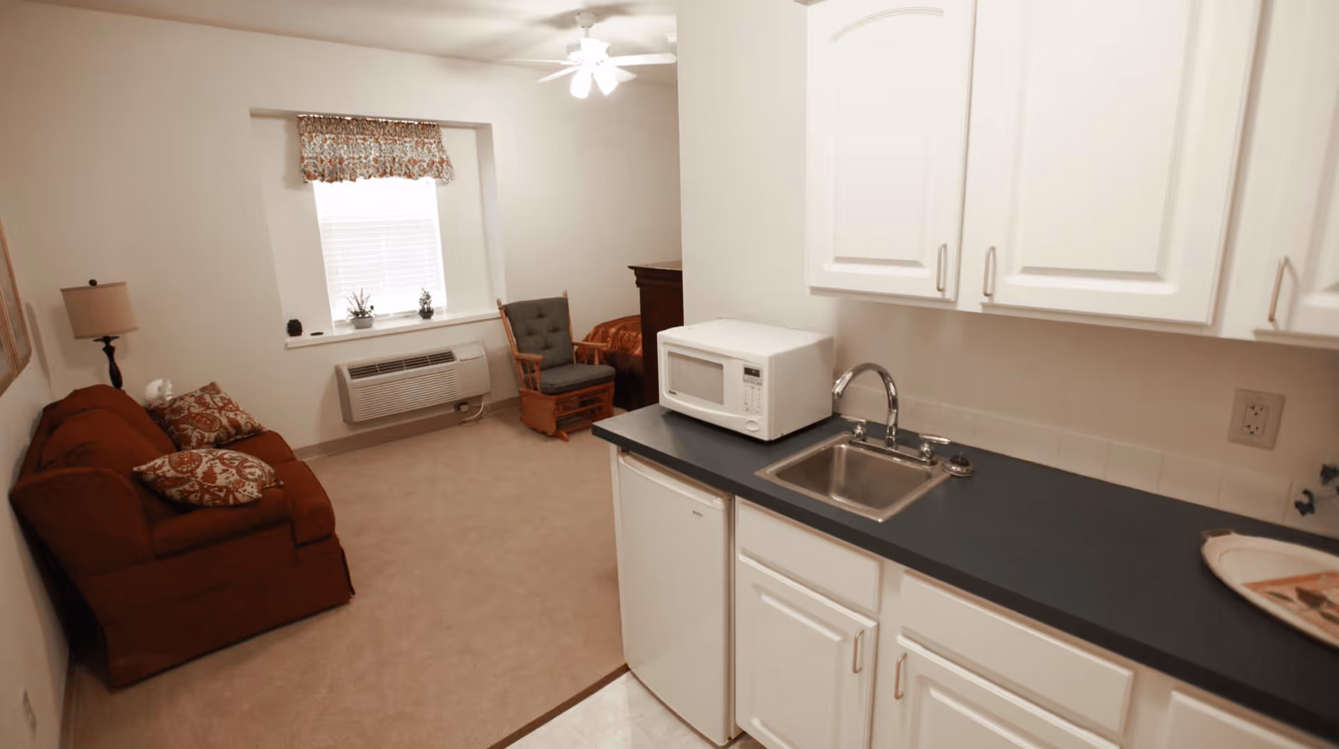 Interior view of a small living space in an assisted living facility showing a kitchenette with white cabinets, a microwave, a small sink, and a mini refrigerator. Adjacent to the kitchenette is a living area with a brown sofa with patterned cushions, a wooden rocking chair, a window with blinds and a floral valance, a lamp, and a bed partially visible in the background.