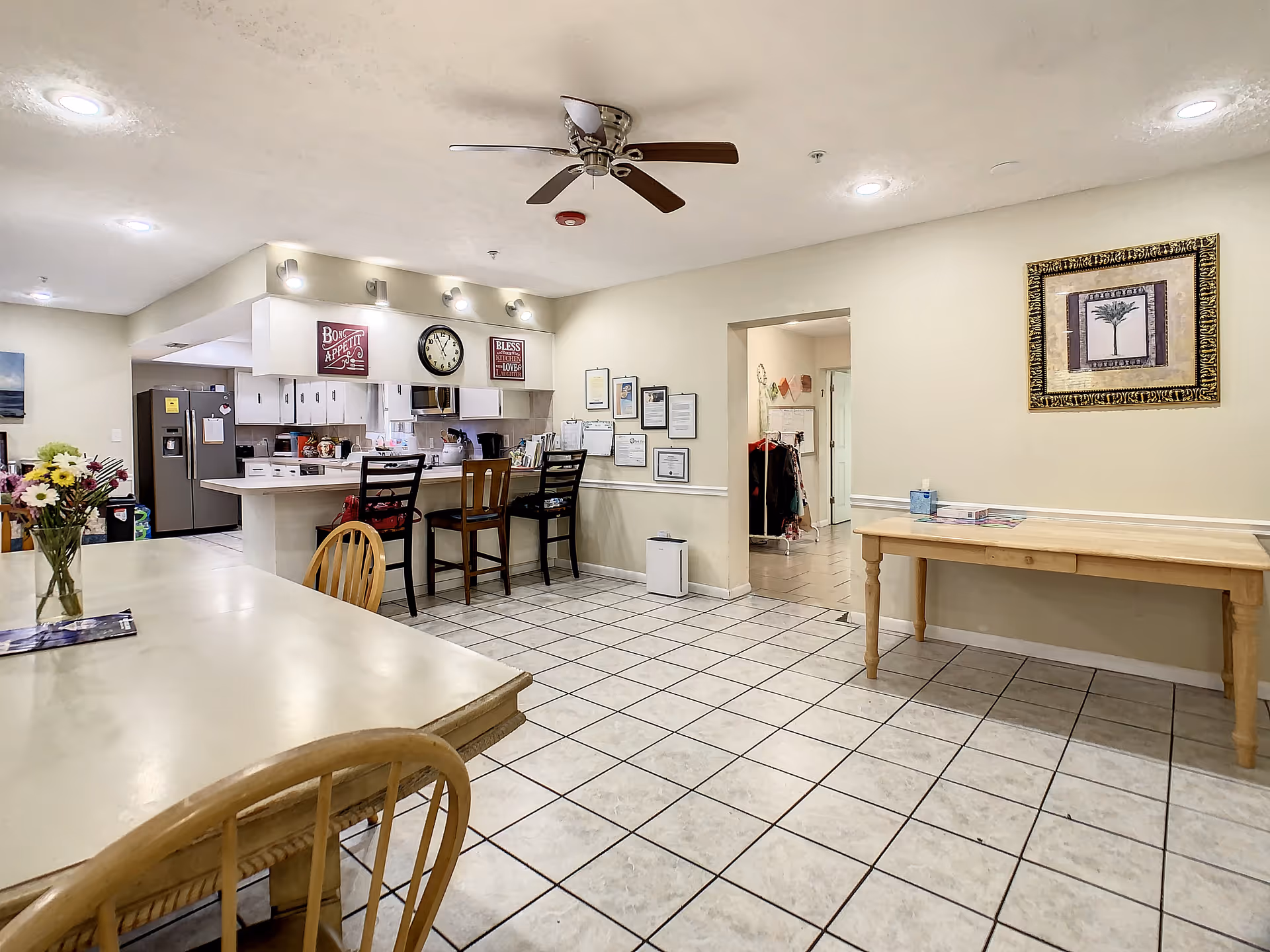 Interior view of a senior living facility dining and kitchen area with tiled floors, a ceiling fan, wooden chairs, a dining table with a vase of flowers, a kitchen counter with bar stools, and wall decorations including a clock and framed pictures.