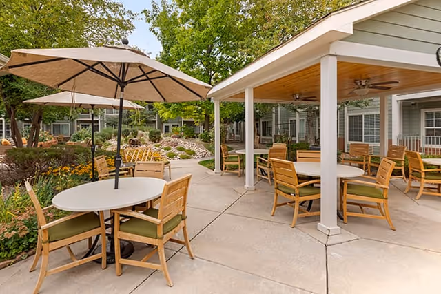 Outdoor patio area at Brookdale Boulder Creek with round tables and wooden chairs featuring green cushions. Some tables have large beige umbrellas. The patio is surrounded by greenery, flowers, and trees, with a covered seating area supported by white pillars and ceiling fans.