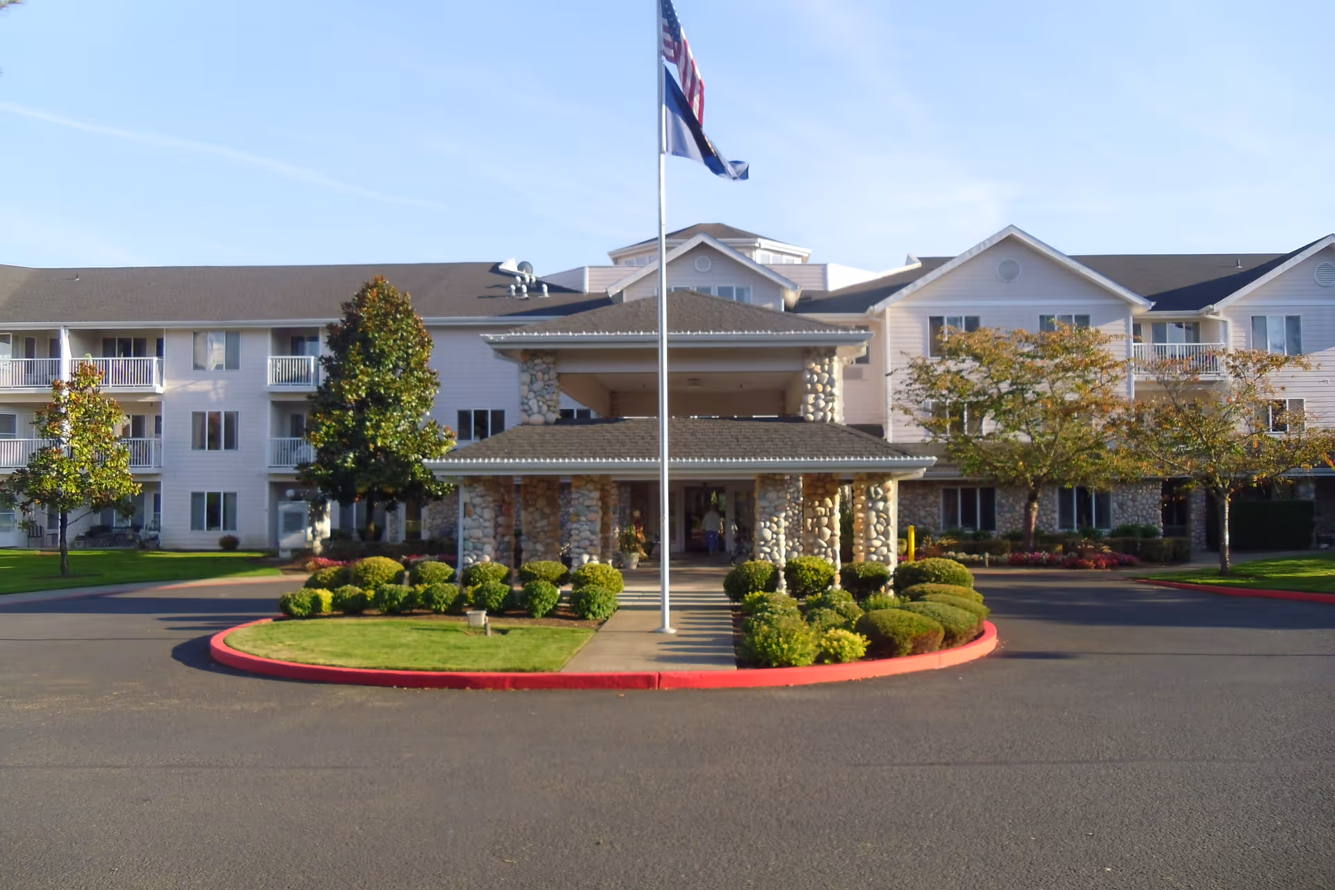 Front exterior view of Somerset Lodge Gracious Retirement Living, showing a three-story building with balconies, a covered entrance supported by stone pillars, a flagpole with two flags, and well-maintained landscaping including bushes and trees.