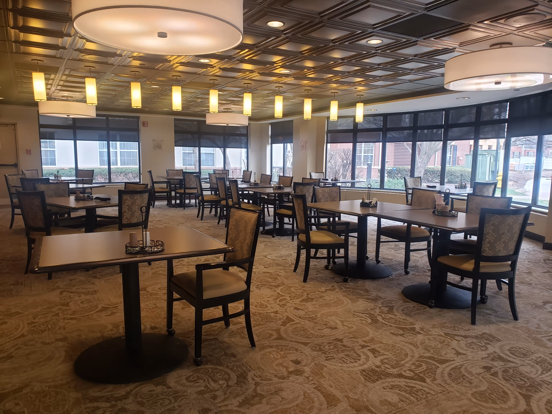 A spacious dining room with multiple square tables and cushioned chairs arranged neatly. The room features large windows with black roller shades, allowing natural light to enter. The ceiling has a decorative pattern with several modern pendant lights and large circular ceiling lights providing warm illumination. The carpet has a subtle patterned design.