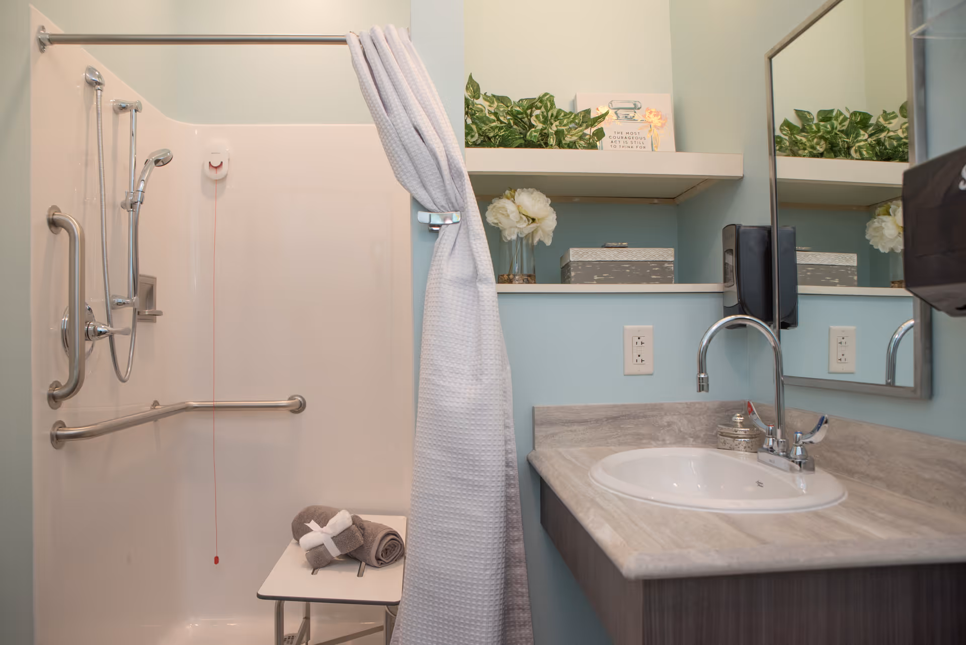 A bathroom with a walk-in shower featuring grab bars and a handheld showerhead. A white shower curtain is partially drawn to the side. Inside the shower, there is a foldable shower seat with rolled towels on it. Next to the shower is a sink with a marble countertop, a faucet, and a mirror above it. On the wall above the sink are two shelves holding decorative plants, a small box, and a framed sign. The walls are painted light blue.