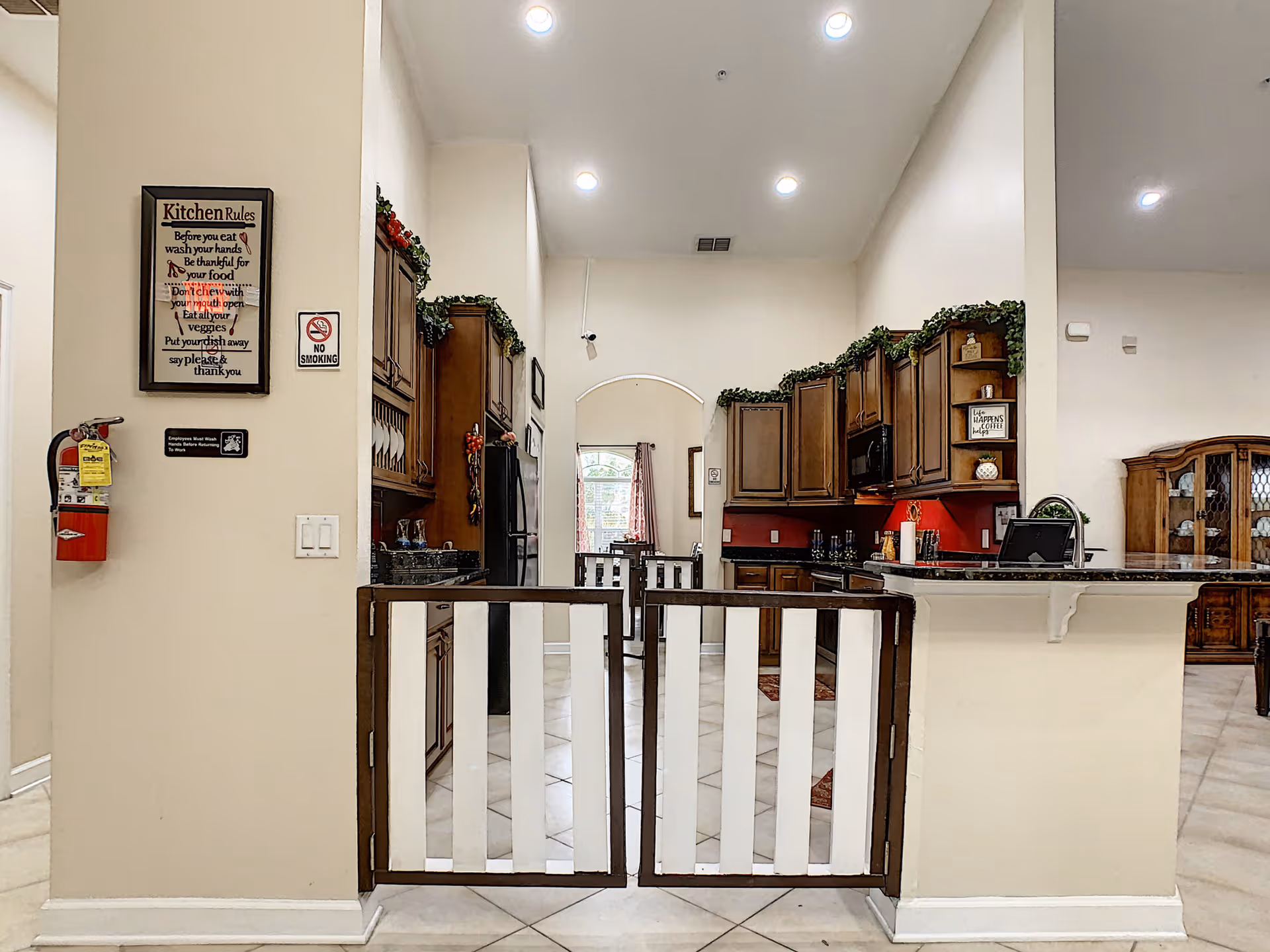 Interior view of a kitchen area in a senior living facility with wooden cabinets, granite countertops, and a tiled floor. A wooden safety gate is installed at the entrance to the kitchen. On the left wall, there are signs including kitchen rules and a no smoking sign, as well as a fire extinguisher. The kitchen is decorated with green garlands on top of the cabinets and has recessed ceiling lights.