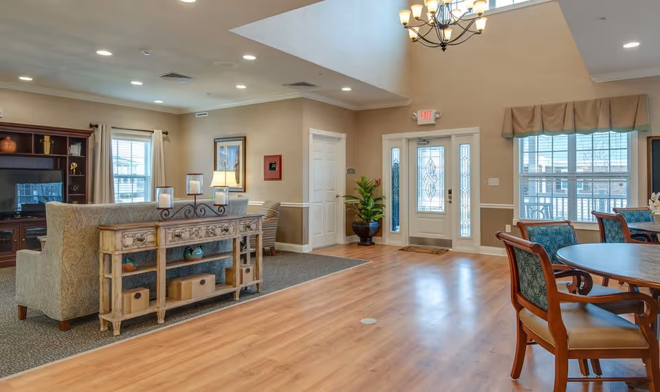A bright and spacious senior living common area with a seating arrangement including a patterned sofa and armchairs, a wooden console table with decorative items, a large window with curtains, a round dining table with chairs, and a glass-paneled front door letting in natural light. The room features wood flooring, beige walls, a chandelier, and a potted plant near the entrance.