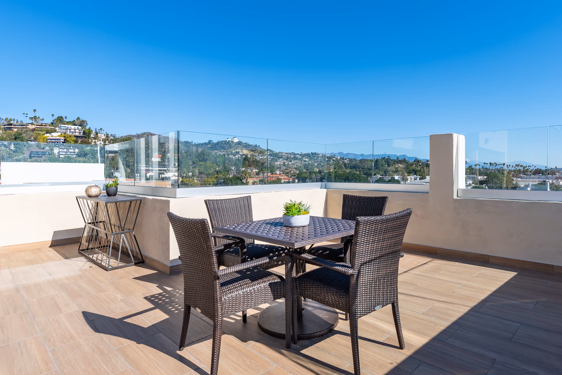 Outdoor patio area with a square table and four wicker chairs, a small side table with decorative items, glass railing, and a clear view of hills and buildings under a bright blue sky.