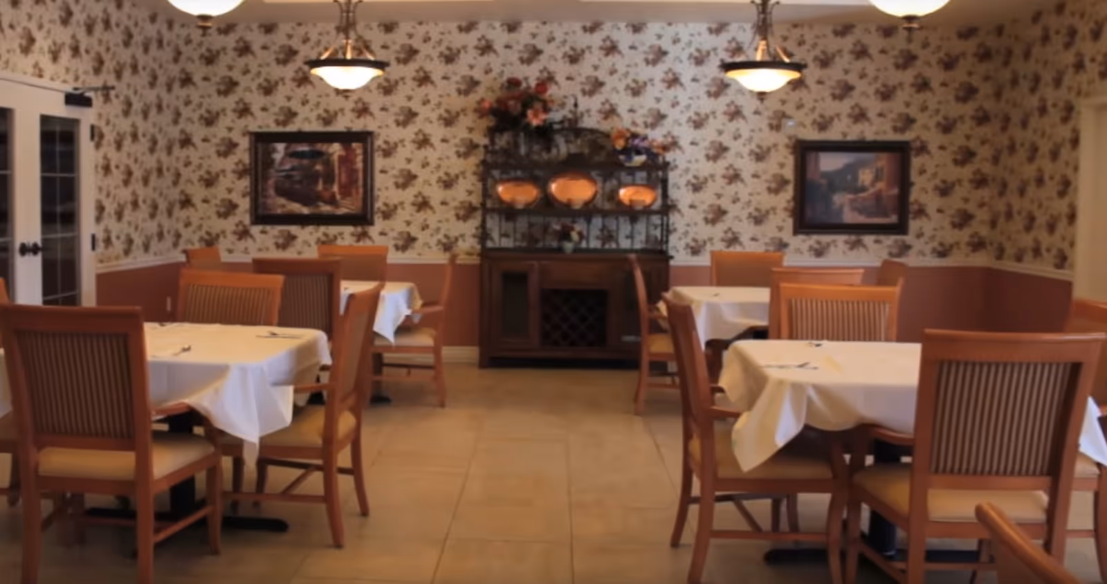 Dining room with several wooden tables and chairs, each table covered with a white tablecloth and set with silverware. The walls have floral wallpaper and two framed paintings. There is a wooden cabinet with decorative plates and flowers in the center of the back wall, and two ceiling lights hang above.
