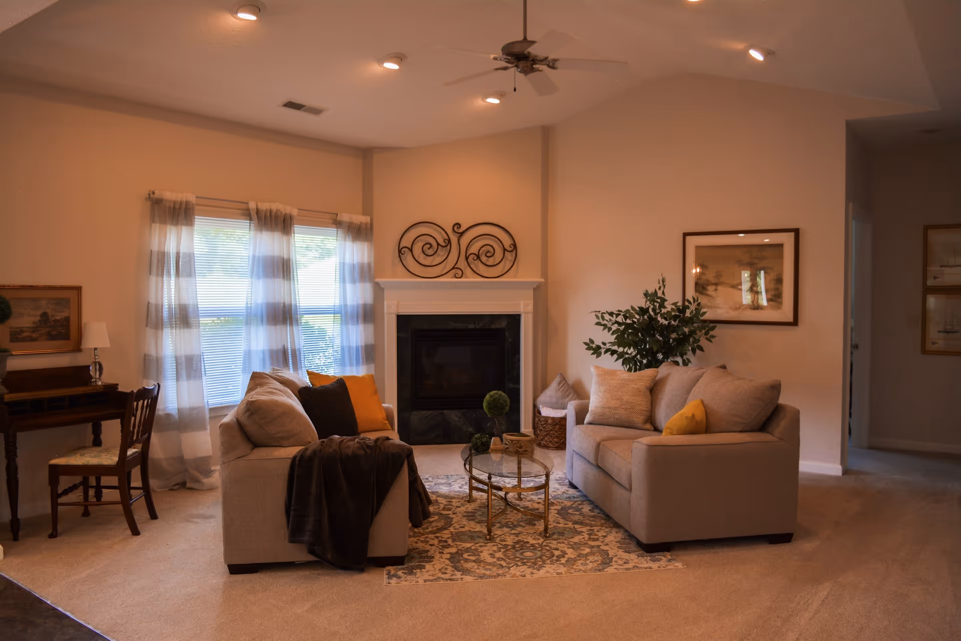 A cozy living room with two beige sofas facing each other, a glass coffee table on a patterned rug, a fireplace with decorative metal art above it, a ceiling fan with lights, and large windows with sheer striped curtains letting in natural light. There is a small wooden desk with a chair and a lamp on the left side, and framed artwork on the walls.