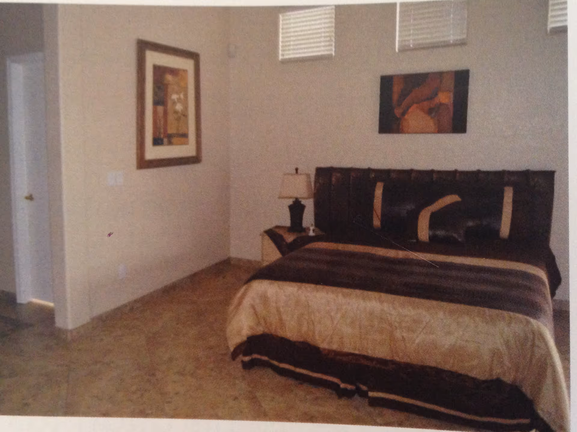 A furnished bedroom with a large bed, dark headboard, nightstand and lamp against beige walls with framed artwork.