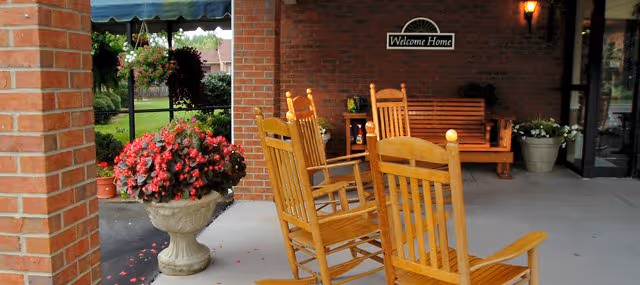 Outdoor covered patio area with wooden rocking chairs and a wooden bench. There is a large planter with red flowers and a hanging basket with greenery. A brick wall with a 'Welcome Home' sign and a lit wall sconce is visible. The area overlooks a green lawn and garden.
