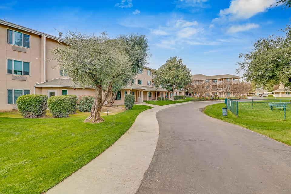 Exterior view of a senior living facility with a paved driveway and sidewalk curving through a green lawn with trees and bushes. The building is three stories tall with beige walls and multiple windows. The sky is partly cloudy with blue patches.