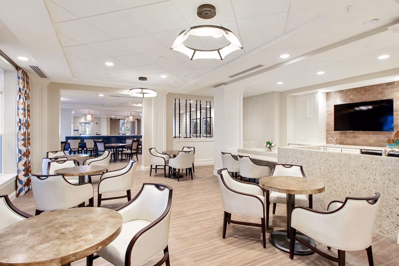 A bright and modern senior living facility common area with multiple round tables and cushioned chairs arranged for seating. The room features wood flooring, large windows with patterned curtains, ceiling lights, and a wall-mounted flat-screen TV above a countertop with a sink. The space is open and inviting, designed for socializing and relaxation.