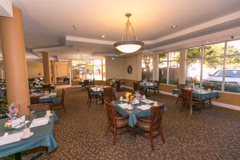A spacious dining room with multiple tables covered in teal tablecloths, each set with cups, glasses, napkins, and small flower arrangements. The room has large windows letting in natural light and a patterned carpet floor. There are ceiling lights and beige walls with decorative elements.