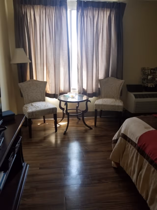 A cozy room with two upholstered chairs and a small round glass table between them in front of a window with closed curtains. To the right is a bed with a beige and red comforter, and to the left is a dark wooden TV stand with a television. There is a floor lamp behind one of the chairs and a heating/cooling unit beneath the window.
