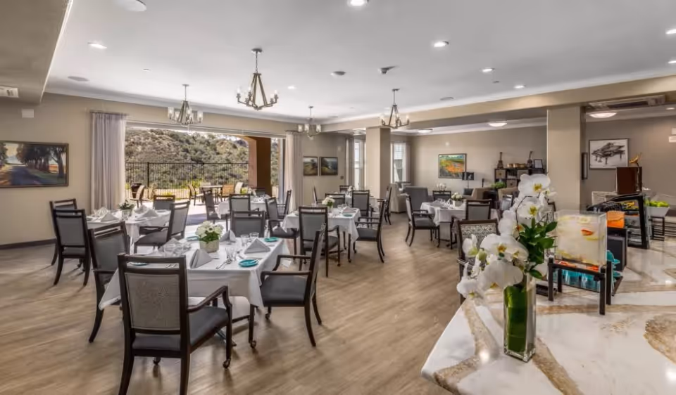 Bright, spacious dining room set with multiple tables and chairs, chandeliers, place settings, and a view out to a patio.