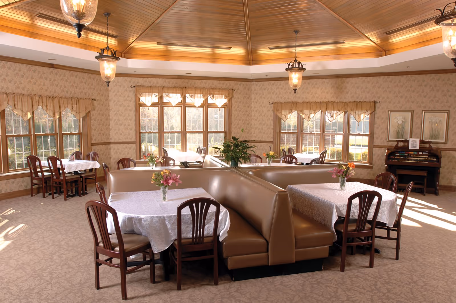 A bright dining room with multiple tables covered in white tablecloths, each adorned with a small vase of flowers. The room features large windows with beige curtains allowing natural light to fill the space. There are brown cushioned booth seats and wooden chairs around the tables. The ceiling is wooden with a geometric design and hanging light fixtures. An organ and framed artwork are visible against one wall.