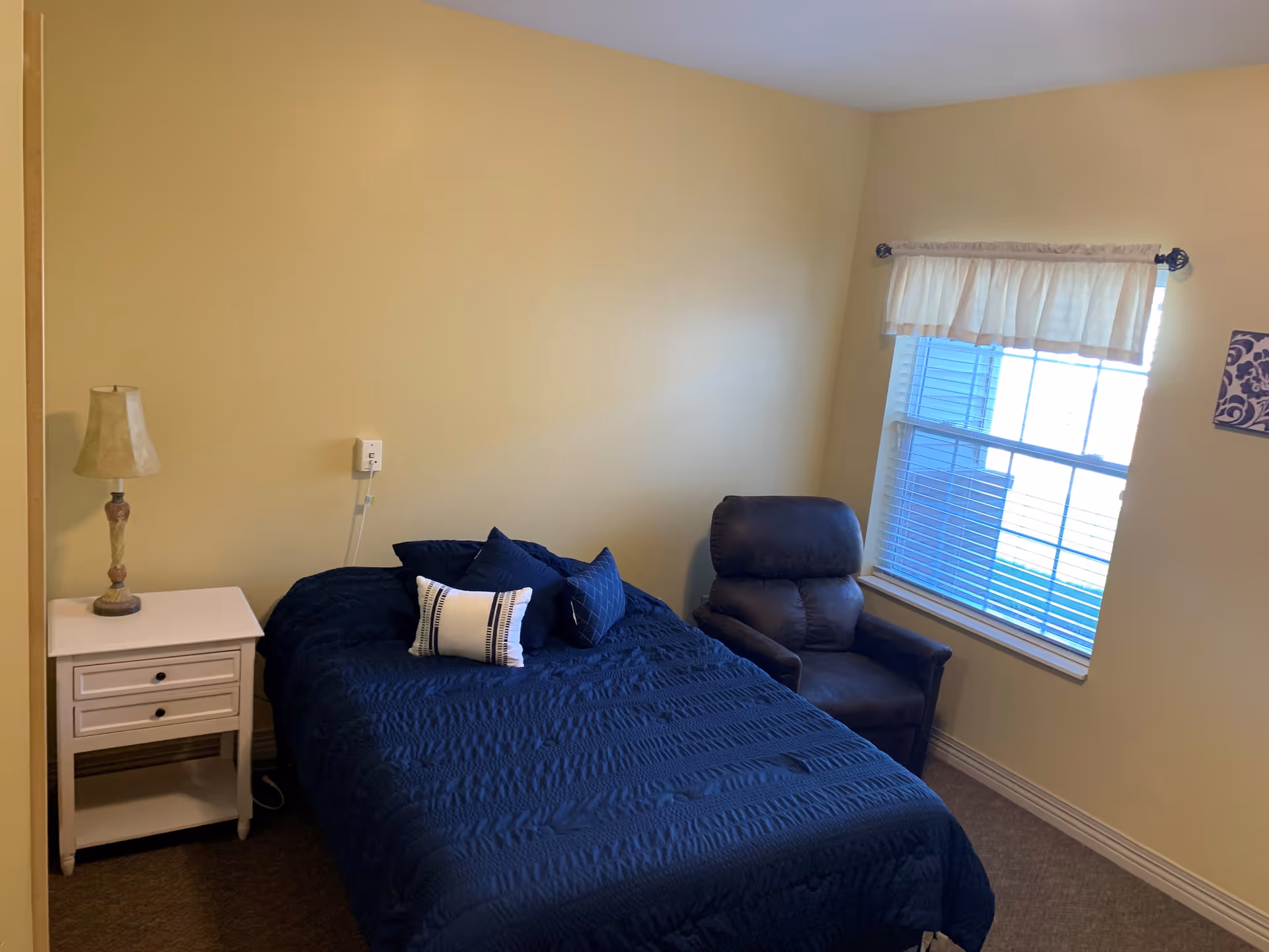 A small bedroom with a bed covered in a dark blue quilt and matching pillows. Next to the bed is a white nightstand with a decorative lamp. A dark brown recliner chair is positioned near a window with white blinds and a short white curtain. The walls are painted light yellow.