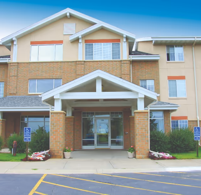 Front entrance of a multi-story senior living building with a covered portico, glass double doors, and accessible parking signs.